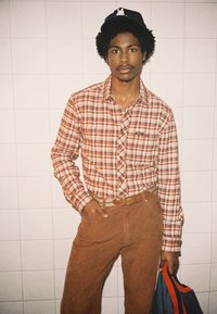 Camicia a quadri in tonalità di arancione e marrone, pantaloni di velluto a coste marroni e un cappellino nero. La persona tiene in mano una borsa blu con dettagli rossi.