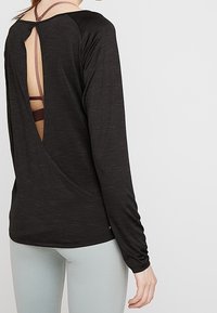Chemise noire à manches longues avec un dos en V qui révèle un soutien-gorge de sport marron foncé en dessous ; fabriquée en tissu doux et texturé.