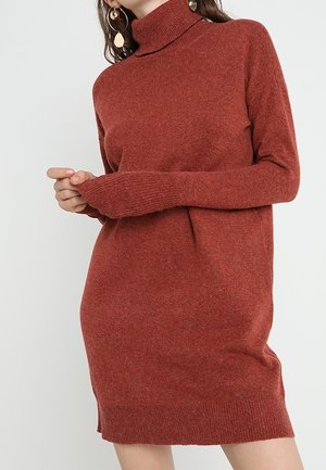 Frau trägt ein rostfarbenes, langärmliges Rollkragen-Strickkleid und hält einen Ärmel nahe ihrer Taille, vor einem weißen einfarbigen Hintergrund.