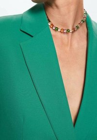 Vihreä bleiseri, jossa on terävät läpät ja syvä V-pääntie, yhdistettynä värikkääseen helmikaulaketjuun, jossa on erilaisia sävyjä ja kuvioita.