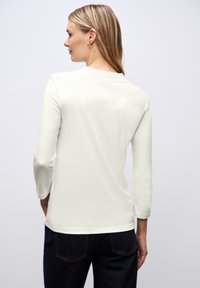 Top de manga larga ajustado en algodón crema, con un cuello redondo y una textura suave. Se lleva con jeans oscuros. Vista trasera mostrada.