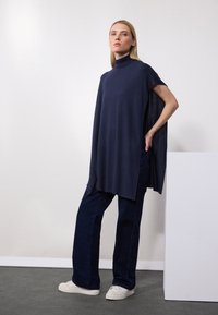 Navy blue tunic with a high neckline, short sleeves, and a loose fit, paired with dark denim trousers and white sneakers.