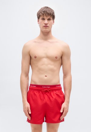 Red swim shorts with drawstring waist, featuring a Calvin Klein logo in white on the waistband. Smooth fabric with a relaxed fit.