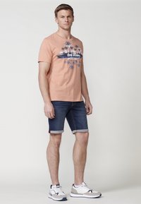 Gai persiku t-krekls ar palmu koka grafiku un tekstu. Apvienots ar tumšiem džinsu šortiem, ar sarullētām piedurknēm, un baltām sporta kurpēm ar tumši zilām akcentiem.