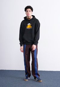 Sort hættebluse med casual logo, brune bukser med blå striber og mørke sneakers. Glat stof, simpelt design, ingen mønstre.
