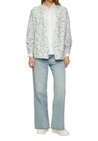 Witte textuur cardigan met donkere spikkels, gedragen over een wit shirt. Gecombineerd met lichtblauwe wijde jeans en witte platform sneakers.