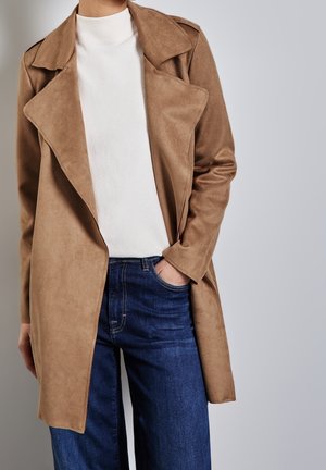 Persoon in een camel-kleurige suède trenchcoat over een witte turtleneck trui en blauwe spijkerbroek, met een hand in de zak.