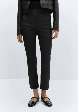 Osoba ubrana w czarne, dopasowane spodnie z wysokim stanem, czarną skórzaną kurtkę oraz czarne lakierowane buty Mary Jane, stojąca na jasnym, jednolitym tle.