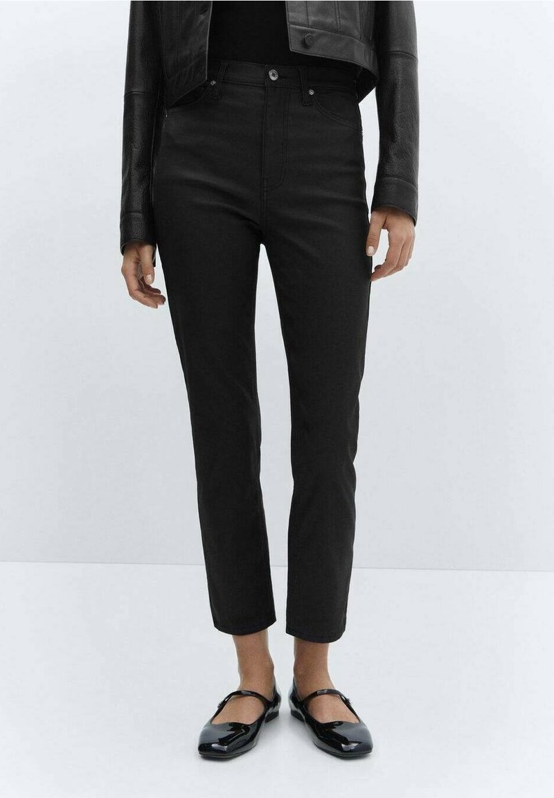 Personne portant un pantalon noir slim taille haute, une veste en cuir noire et des chaussures Mary Jane en cuir verni noires, debout sur un fond clair uni.
