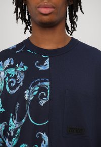 Mornarska majica s stranskim panelom z zapletenimi modrimi cvetličnimi vzorci, običajno žepom in oznako Versace Jeans Couture.