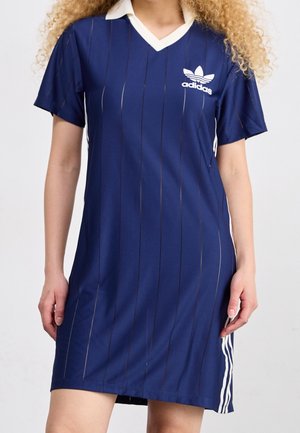 Vrouw draagt een blauwe Adidas jurk met korte mouwen, een witte V-halskraag en drie witte strepen aan de zijkant.