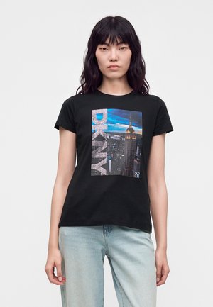 Femme aux cheveux noirs portant un t-shirt noir avec une vue panoramique de New York et le logo DKNY, associée à un jean bleu clair, debout devant un fond uni.