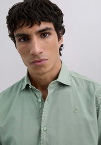 Joven con cabello oscuro y ojos marrones, vistiendo una camisa de botón color verde claro con un pequeño logo de calavera en el pecho, fondo neutro.