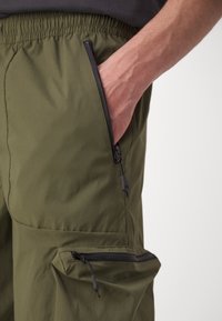 Main dans la poche d'un pantalon vert olive avec des fermetures éclair noires visibles sur le côté et une poche cargo, sur un fond uni.