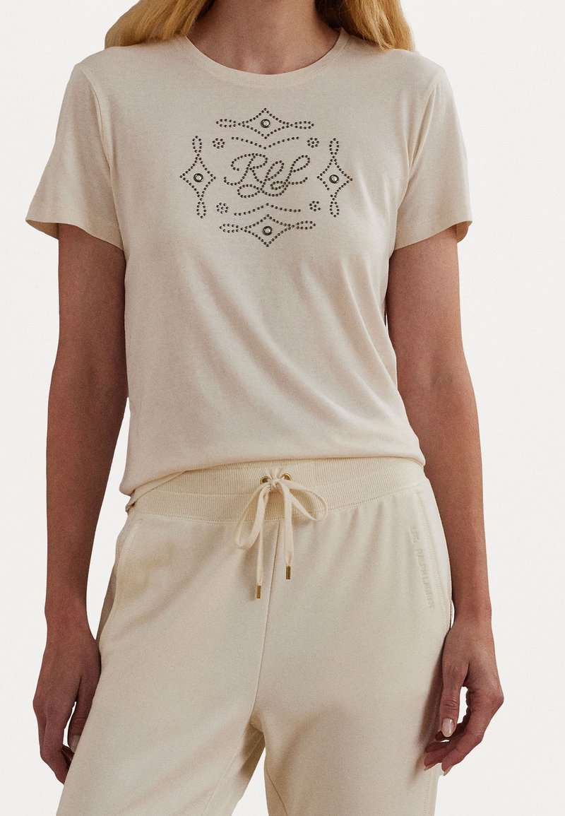 Beige t-shirt med dekorative rhinestones, der danner et mønster. Blødt, glat stof med korte ærmer, parret med lyse bukser med snøre.