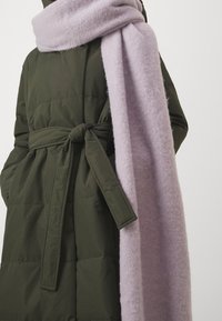 Olivově zelená prošívaná bunda s uvázaným páskem, doplněná jemným světle fialovým šátkem. Šátek má nadýchanou texturu a elegantně splývá.