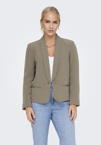 Blazer taillé vert olive avec revers pointus et fermeture à un bouton, associé à un top blanc et un jean bleu. Texture de tissu lisse.