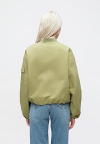 Olivenfarget bomberjakke, lett materiale, elastiske ermer og nederkant, lomme på venstre erme, brukt over blå jeans.