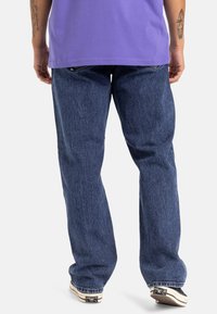 Pantalones vaqueros de mezclilla azul con un corte relajado, que presentan una textura sutil, diseño de cinco bolsillos y un ligero desgaste, combinados con zapatos negros.
