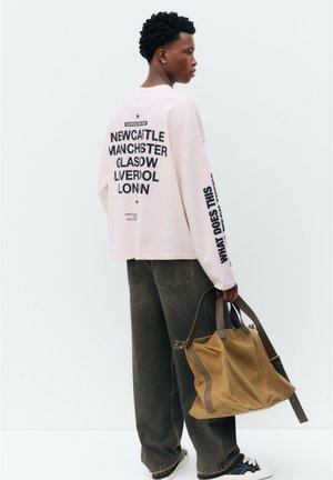 Jeune personne portant une chemise blanche à manches longues avec des noms de villes imprimés dans le dos, tenant un grand sac en daim beige, portant un jean foncé et des baskets.
