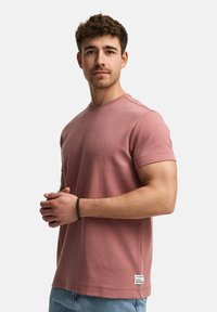 Camiseta de algodón en rosa empolvado con mangas cortas, cuello redondo y detalle de etiqueta lateral. Combinada con pantalones cortos de denim azul claro.