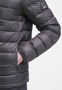 Chaqueta negra acolchada con diseño acolchado, que presenta un parche de logo en la manga y un ajuste ajustable. El material parece brillante y suave.