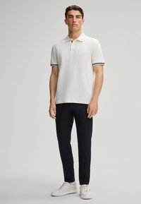 Biała koszulka polo z krótkimi rękawami, teksturowany materiał, akcenty w czarno-białe paski na rękawach, w połączeniu z ciemnymi spodniami i białymi trampkami.