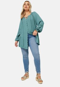 Turkos oversize blus med veck, knäppning och långa ärmar; matchad med ljusblå skinny jeans och kilklacksandaler.