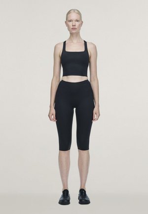 Une femme se tient face à l’objectif devant un arrière-plan uni. Elle porte un débardeur court noir, un legging arrivant aux genoux et des baskets.