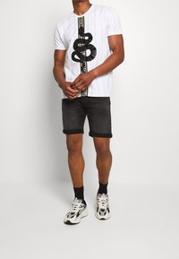 Vit t-shirt med kort ärm och horisontella ränder, med en svart ormgrupp och guldaccenter. Parat med svarta denim-shorts och sneakers.