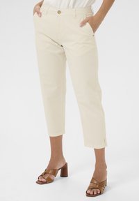 Helle beigefarbene, cropped Hose mit zwei seitlichen Taschen, einem vorne liegenden Knopfverschluss und umgeschlagenen Säumen. Kombiniert mit braunen Riemchensandalen mit Absatz.