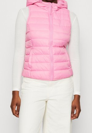 Kobieta ubrana w bezrękawnik różowy pikowany z frontowym zamkiem błyskawicznym, na białej koszuli z długim rękawem i białych spodniach, stojąca z rozluźnionymi ramionami.