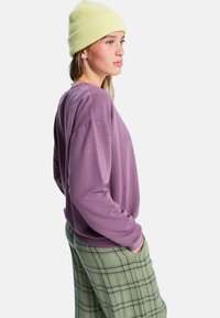 Sudadera de color púrpura con costuras en rosa contrastantes, combinada con pantalones de cuadros verdes. El modelo lleva un gorro amarillo de canalé.