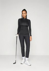 Ženska v črni golf obleki drži golf palico, stoji pred enostavnim svetlim ozadjem, nosi bele rokavice in bele superge.