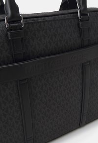 Sac fourre-tout noir avec un tissu texturé arborant un motif logo discret, agrémenté d'accents en cuir lisse et d'un nom de marque en relief.