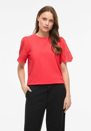 Femme aux longs cheveux bruns portant un chemisier rouge à manches courtes avec des détails en dentelle et un pantalon noir, debout avec une main dans la poche.