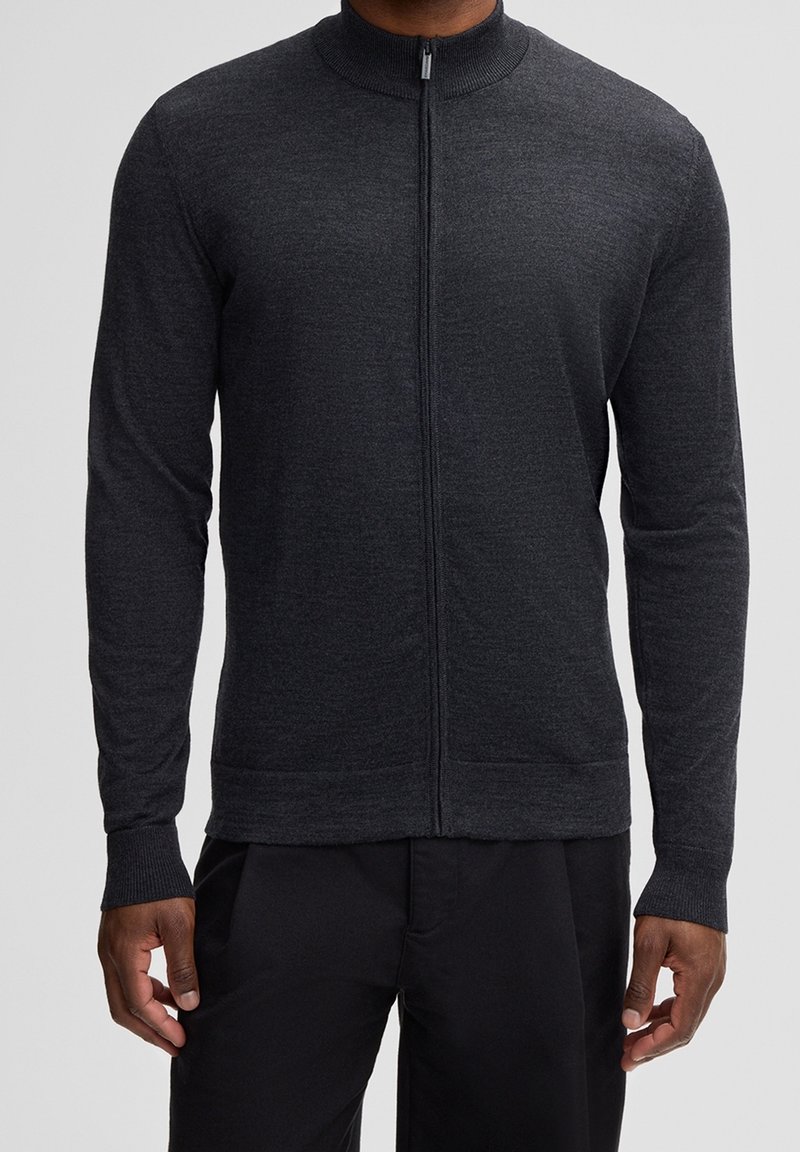 Homme portant un pull gris foncé à manches longues, à fermeture éclair complète, avec un col et des poignets côtelés, associé à un pantalon noir.