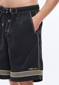 Μαύρα μαγιό βερμούδα με κορδόνι στη μέση, κίτρινες λεπτομέρειες, πλαϊνές τσέπες και γκρι περιθώριο στο τελείωμα. Το υλικό έχει λεία υφή.