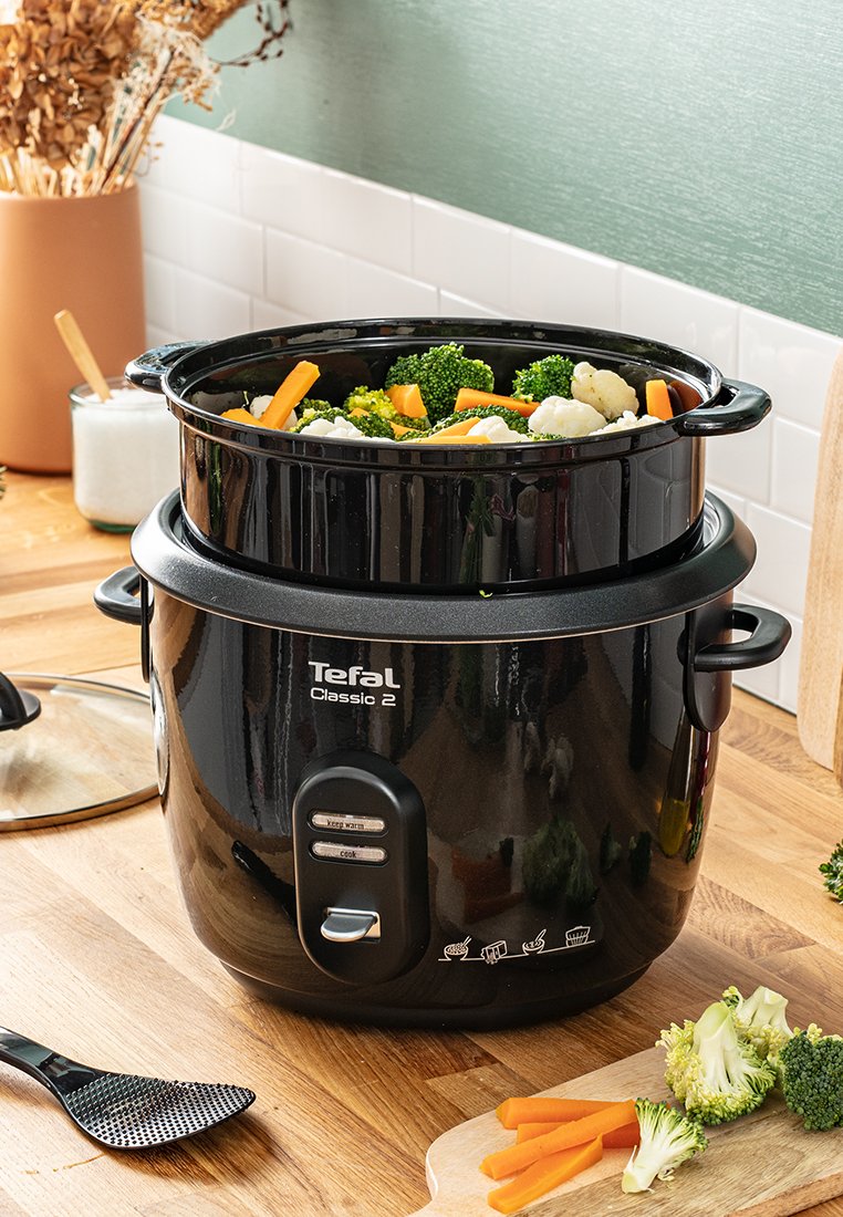 Zwarte rijstkoker met een doorzichtige deksel, gevuld met broccoli, wortels en bloemkool. Beschikt over bedieningsknoppen aan de voorkant en zijhandvatten.