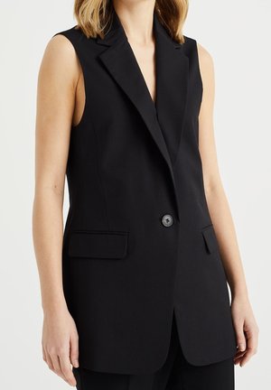 Kvinde iført sort ærmeløs blazer med en knap og klaplommer, vist fra midt på brystet til hofterne mod en ensfarvet baggrund.