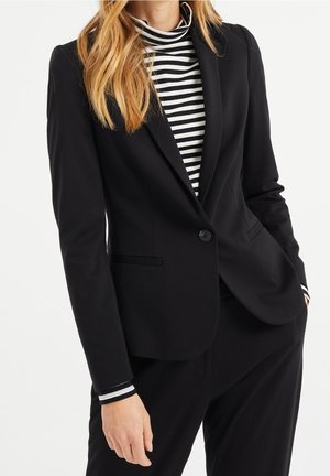 Vrouw draagt een zwarte blazer met één knoop over een zwart-wit gestreepte coltrui met bijpassende gestreepte manchetten, poseert met een hand in de zak.