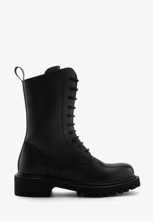 Botín de piel negra hasta el tobillo con puntera redonda, cierre de cordones en la parte delantera y suela de goma texturizada. Cuenta con un tirador en el talón para facilitar su puesta.