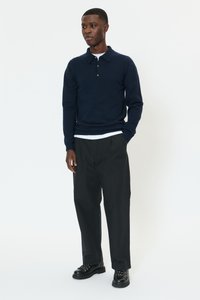 Mörkblå pikétröja med lång ärm och krage, lager-på-lager över en vit skjorta. Kombinerad med svarta byxor och svarta snörskor. Enkel design, utan mönster.