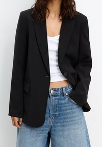 Blazer noir ajusté avec une fermeture à un seul bouton, des revers crantés et un tissu texturé, associé à un débardeur blanc et un jean bleu clair.