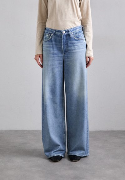 Jean en denim bleu clair à jambes larges avec une teinture délavée, poches avant et fermeture à bouton. Associé à un haut beige à manches longues.
