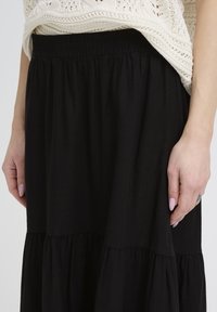 Gros plan sur des mains reposant sur les côtés, portant une jupe noire froncée et un haut en maille crème.