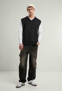 Dark gray sleeveless sweater, white long-sleeve shirt underneath, olive cargo pants with pockets, and white/gray sneakers. Beanie hat.