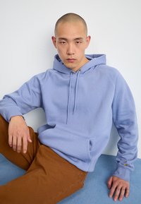 Haut de hoodie bleu clair avec une poche avant et un cordon de serrage, associé à un pantalon marron. Texture douce et design décontracté.