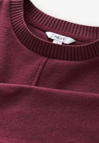 Bordowy sweatshirt z prążkowanym okrągłym dekoltem, gładką fakturą i centralnym szwem. Metka wskazuje rozmiar S, marka NEXT.