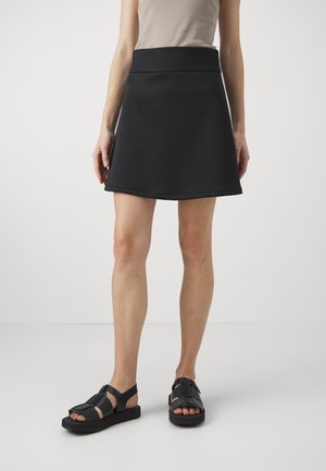 Žena ve černé sukni ke kolenům, béžovém tílku bez rukávů a černých sandálech s odkrytými prsty, stojící na jednobarevném bílém pozadí.